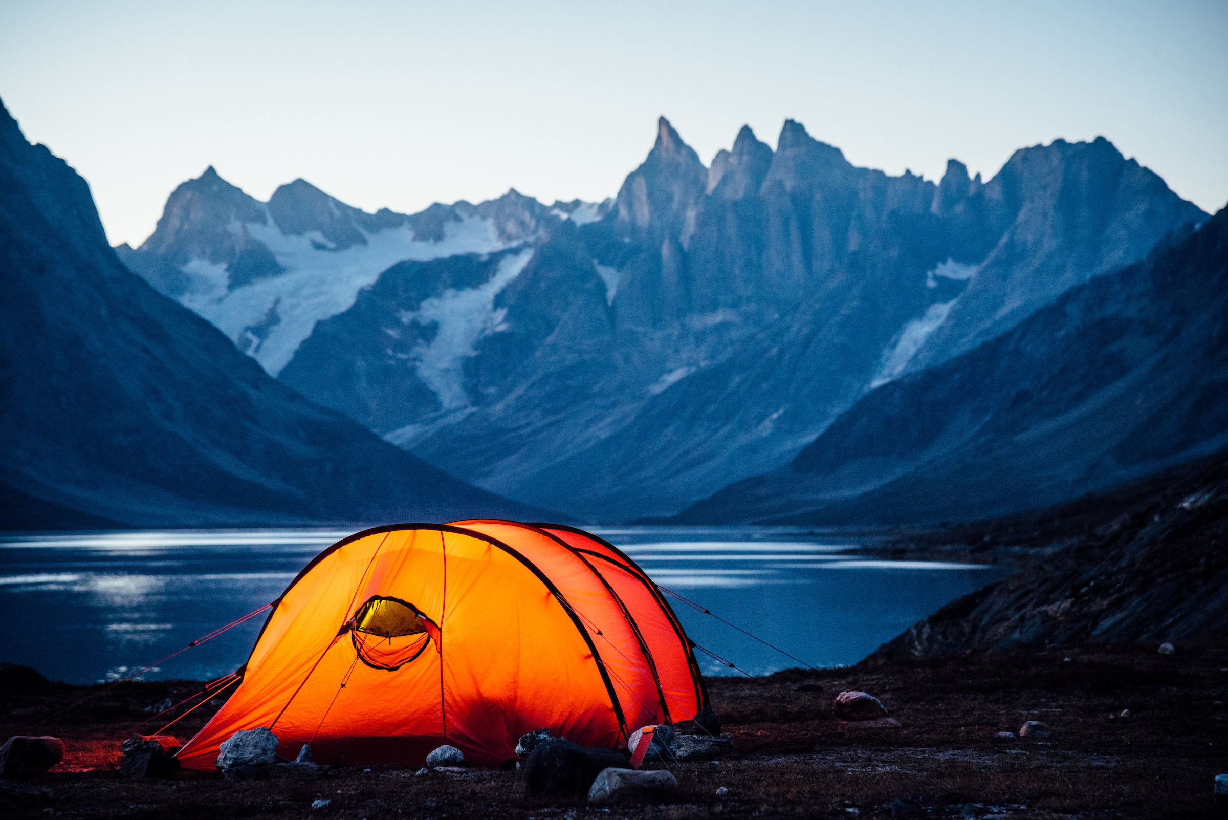 A lit tent at sunset looking north towards three peaks, Camp In Tasiilaq Fjord. Photo by Chris Brin Lee Jr. - Visit Greenland